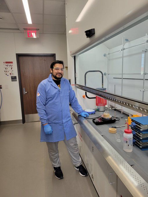 Image of Mudassir standing in the research lab, next to a hood with his first reaction running.