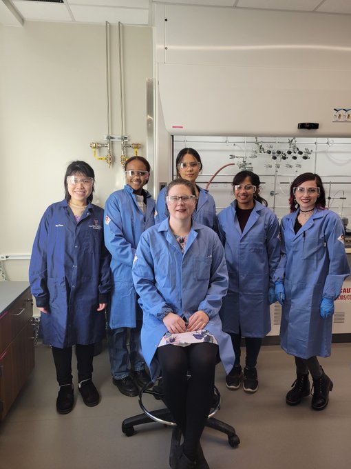 Image of the female members of the Kristufek lab taken in the lab. Professor Kristufek sitting in the front row and five other female members standing in the back. All wearing blue fire-resistant lab coats and safety goggles.